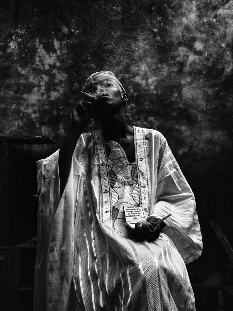 A woman wearing traditional masculine attire smokes a cigarette and sips on dogoyaro, a popular drink made of mixed liquors. Wearing the agbaba and cap and indulging in the traditionally ‘masculine’ habits of smoking and drinking.