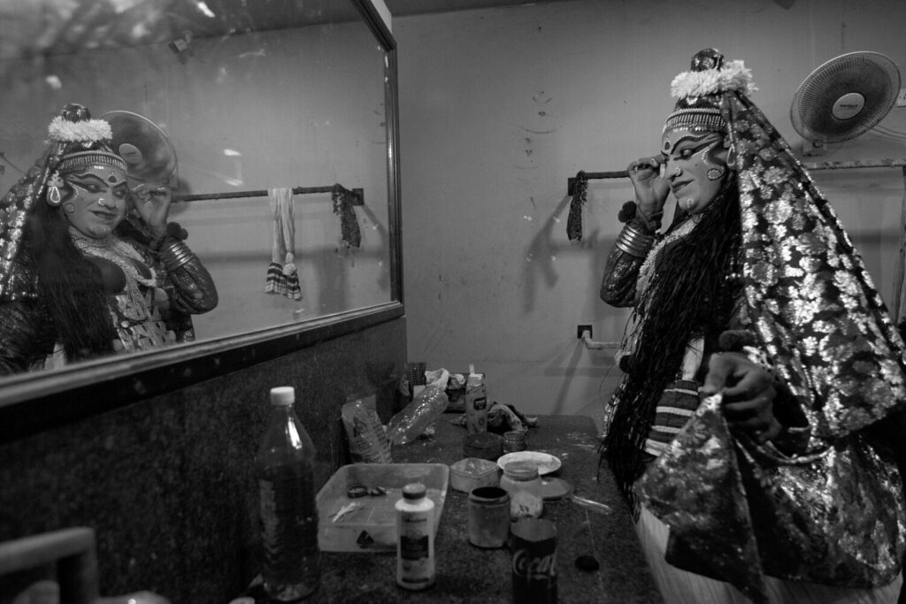 A male actor in his dressing room prepares for his role in the Kathakali dance. Adorned in an elaborate costume and applying thick, stylised make-up, the figure encapsulates a different kind of gender expression.