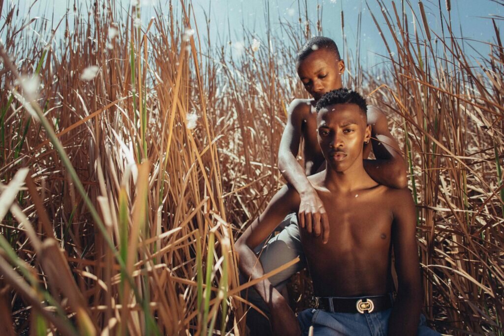 Two non-binary people pose in a field in Cape Town bathed in a warm golden light. The scene is both familiar and other-wordly. Around the figures, delicate balls of cotton ethereally float in the saturated sky and among the long reeds.