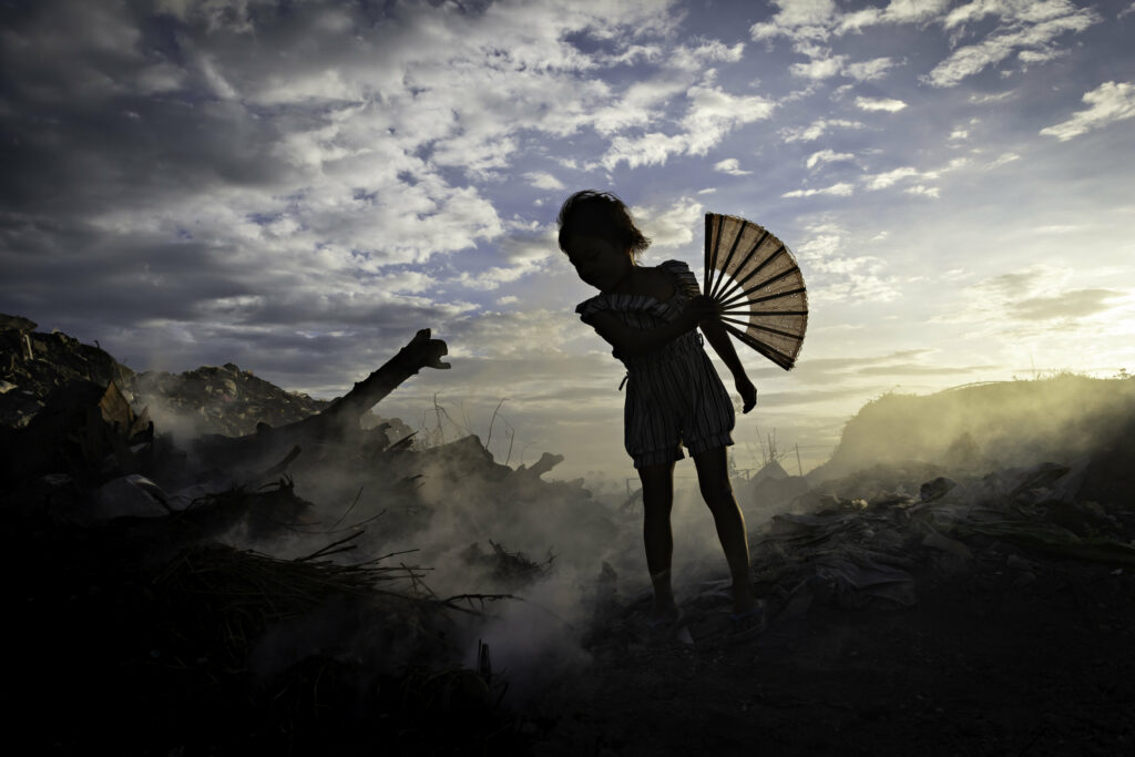 A young girl flourishes a fan above the smouldering wreckage of the Paranaque City landfill. Her small figure, frilly clothes and ornamental fan silhouetted against the darkening sky dramatically frames the uncomfortable reality of the landfill