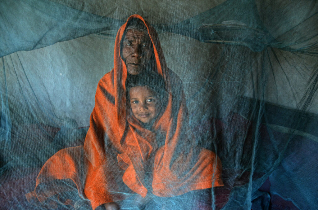 Behind the translucent blue of the mosquito net, grandfather Chandan Chetri swaddles his young grandson. Shrouded in the orange fabric, the boundaries between the bodies of the grandfather and grandson are blurred, and we’re encouraged to meet their direct gaze