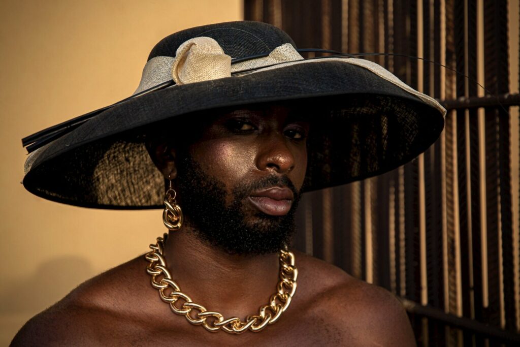 Dressed in a Sunday hat and adorned with heavy gold jewellery, a man poses for the camera. His face, knowingly turned towards the lens shimmers with golden highlighter and blush.