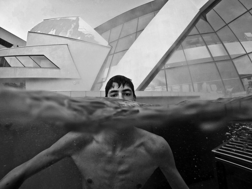 As the water bends the light, the boy’s submerged body appears enlarged in contrast with the upper part of the head that surfaces above the water. Suspended in movement, his slim arms and angular bones uncannily mirror the geometric and imposing building in the background as though he were attempting to imitate the world that surrounds him.