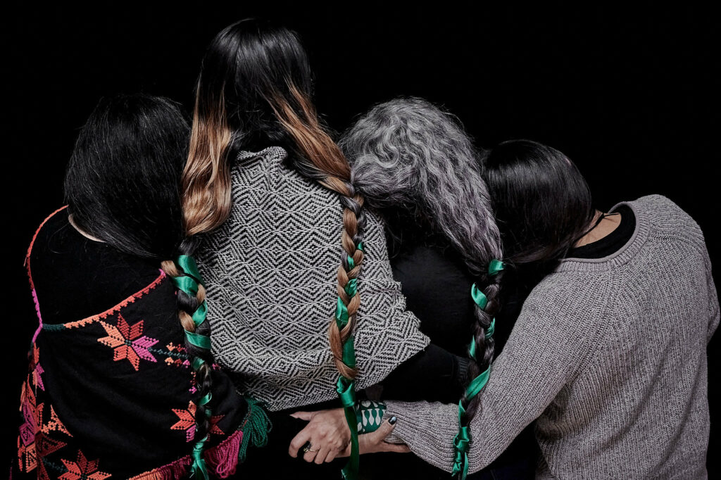 Four people facing away from the camera huddle together with their hair braided and their arms entwined.