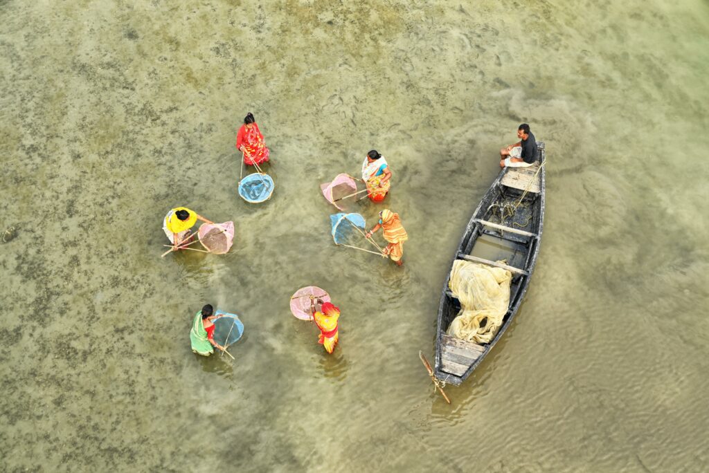 Captured from above, six women clothed in bright fabrics fish with nets in a circle. Their cheerful clothing sets them apart from the muddy flats of low tide and the man in the boat who watches over them.