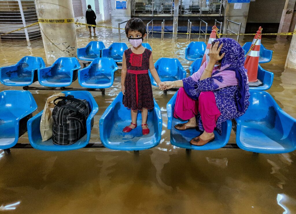 In the flooded lobby of Chittagong Maternal & Childcare Hospital in Bangladesh, a mother and her child, wait to receive medical treatment. They sit and stand on blue plastic chairs that appear to hover over the swirling, muddy water.