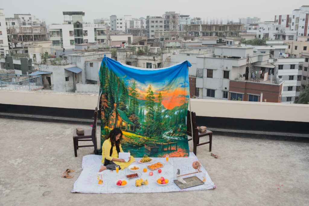 The grey skyline of high-rise buildings contrasts with a colourful staged setting depicting an enchanting forest, flowing river, and serene hill. This sombre juxtaposition reflects the diminishing green spaces in Dhaka city due to rapid urbanisation.