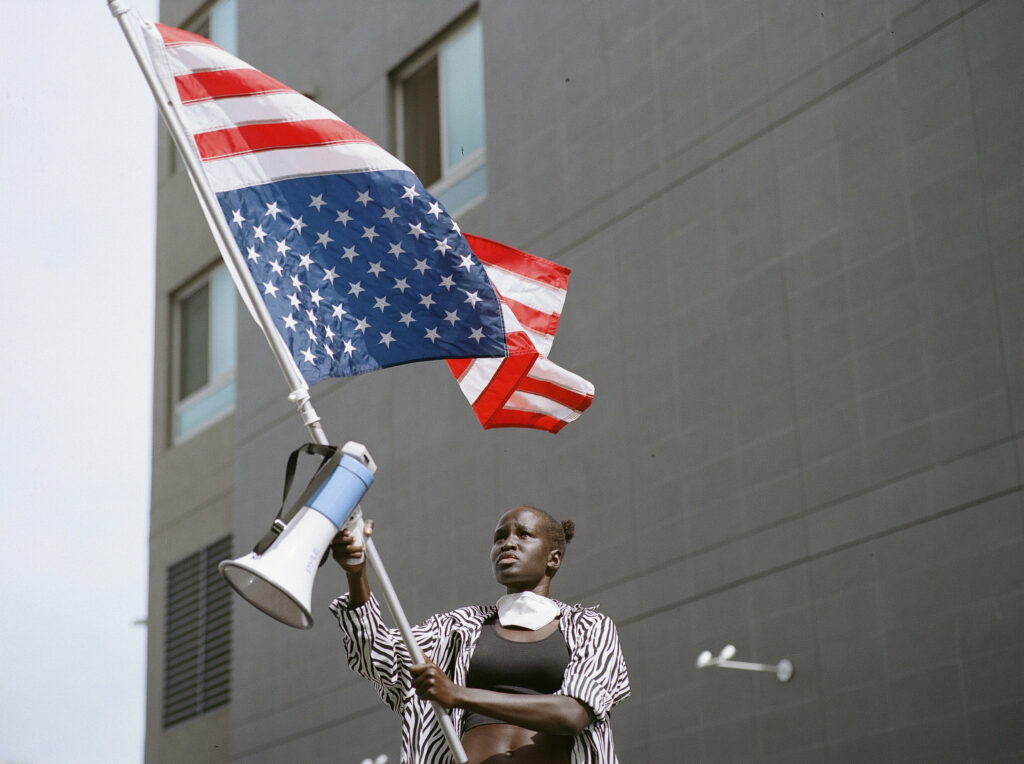 A young girl waves the US flag in front of a large grey building at a Black Lives Matter protest in Los Angeles.