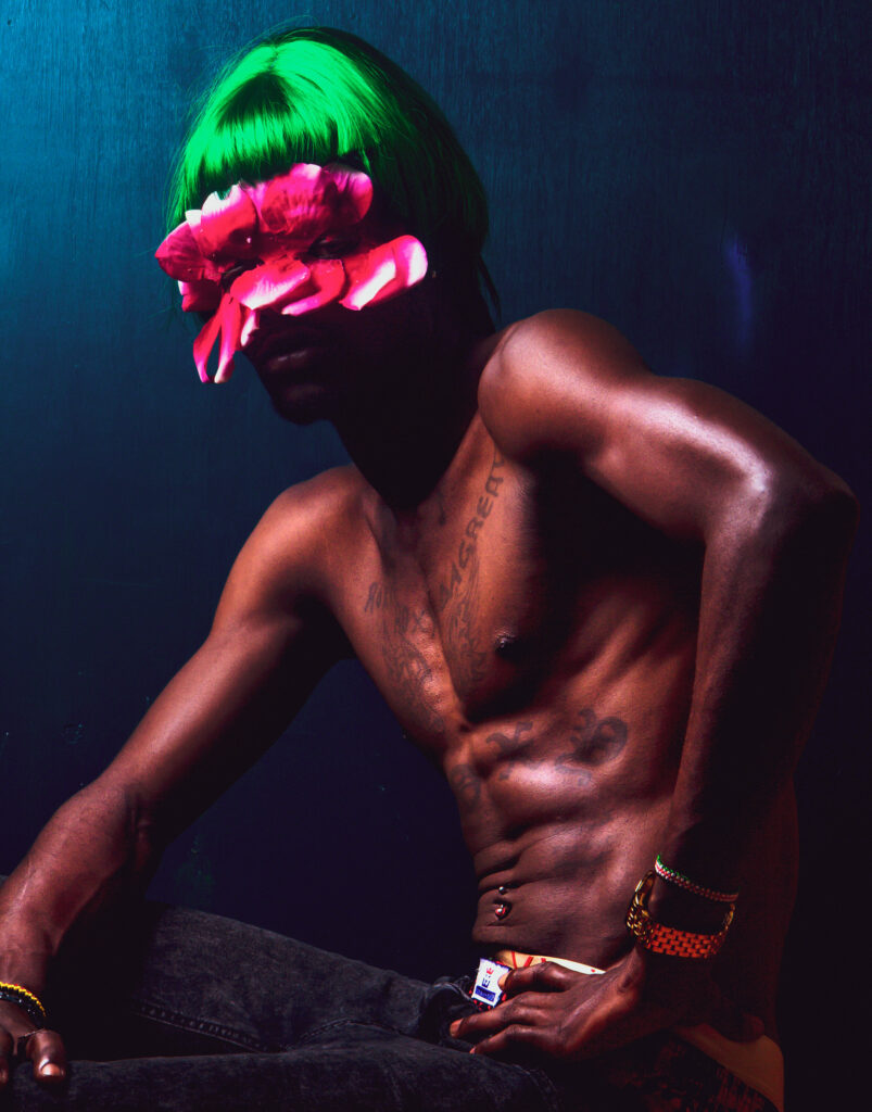 A shirtless man poses in a luminous green wig and a pink petal mask that obscures his eyes.