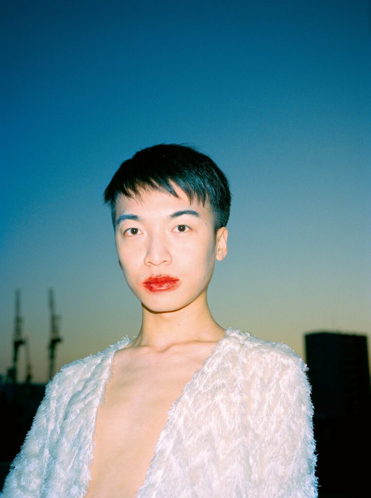 A young man stares directly into the camera in front of a blurry blue backdrop, his red lipstick slightly smeared.