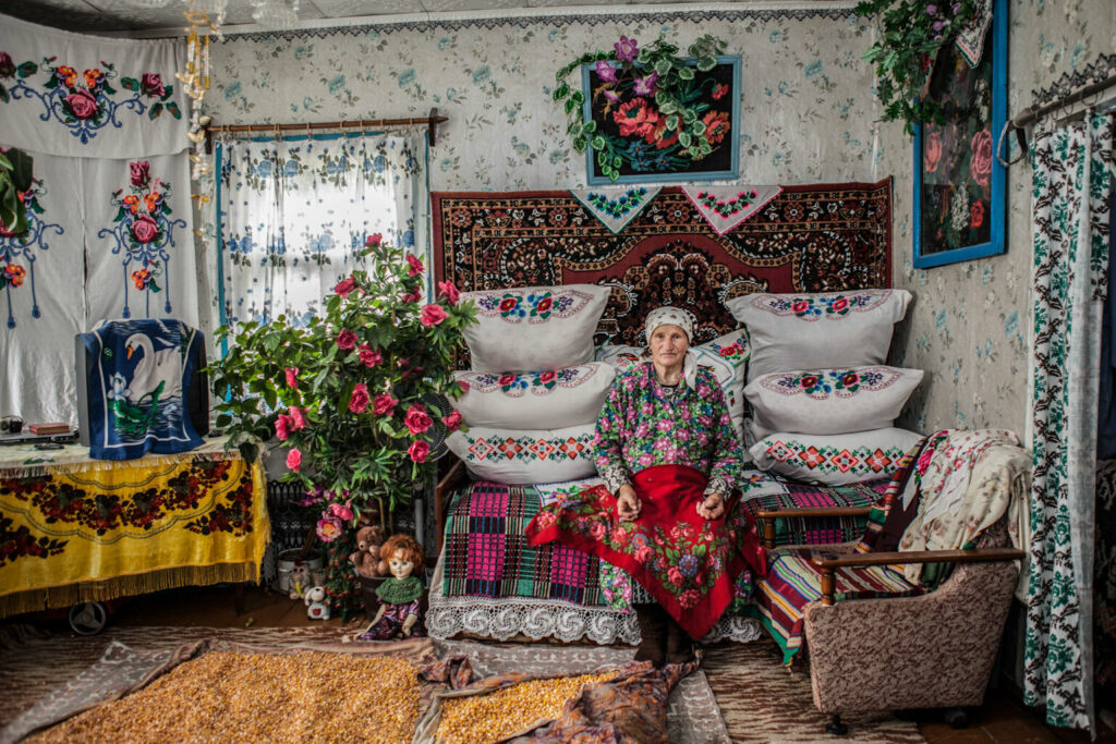 A widowed woman sits at her home in Belarus. Surrounded by flowers, embroidery and teddy bears, she dries grain on her carpeted floor.