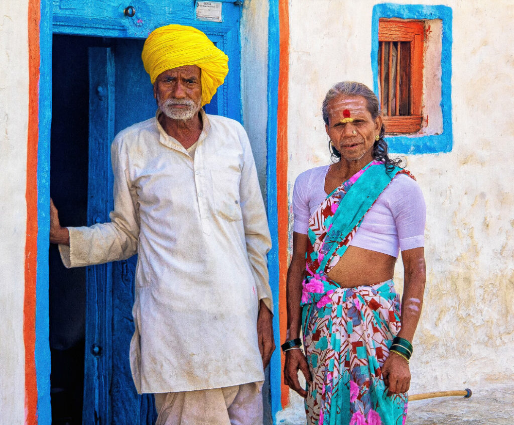 A husband and his transgender wife stand in front of the home that they have shared for 40 years in Karnataka, India. In front of their vibrant home, the couple stare out towards us. Their stance is defiant as they pose squarely towards the camera.