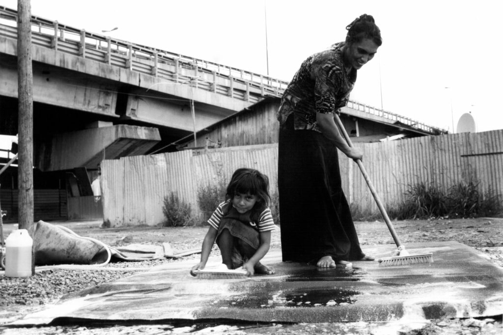 Hunched over the broom, a woman scrubs a mat. Next to her, a young girl squats, barefooted, scrubbing the soapy water. In the cold, corrugated setting, a warmth radiates from the two figures, who smile awkwardly as they clean.