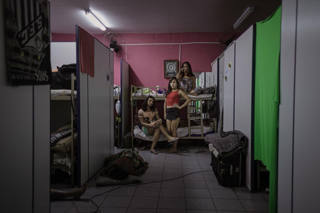 At the end of a dimly lit walkway bordered by makeshift room dividers, Kimberli, Allana and Janaina pose on a bunk bed backgrounded by a pink wall.